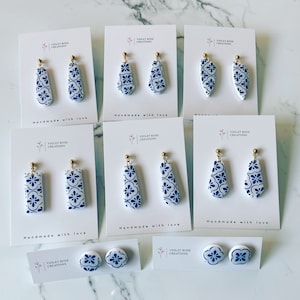 Puede incluir: Un conjunto de pendientes de cerámica con estampado azul y blanco con ganchos dorados. Los pendientes se muestran en tarjetas blancas con el texto "Violet Rose Creations" y "Handmade with love".