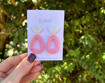 May include: A pair of handmade earrings with a cloud-shaped top and a pink, oval-shaped bottom. The earrings are on a white card with the text "VIOLET ROSE CREATIONS" and "handmade with love". The background is a blurred green.