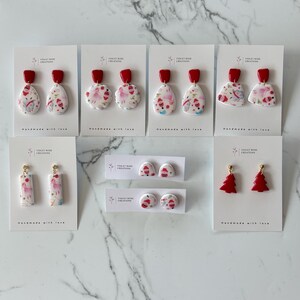 May include: A selection of handmade Christmas earrings featuring various designs, including a red and white Santa Claus, a red and white Christmas tree, and a red and white snowflake. The earrings are displayed on white cards with the text "Violet Rose Creations" and "Handmade with love".