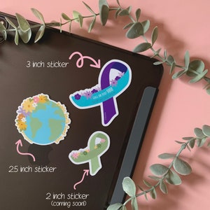 May include: Three different sticker designs featuring a green ribbon with flowers, a blue ribbon with flowers and the text "CALL OR TEXT 988 SUICIDE AND CRISIS LIFELINE", and a blue and green globe with flowers. The stickers are on a black surface.