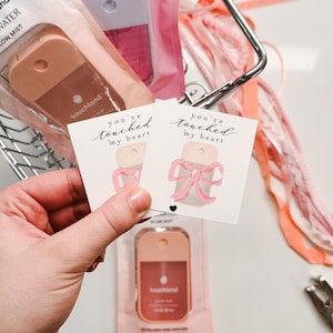 May include: Two small pink and white gift tags with the text "You've touched my heart" and a watercolor illustration of a pink bow. The tags are in a hand holding them up.