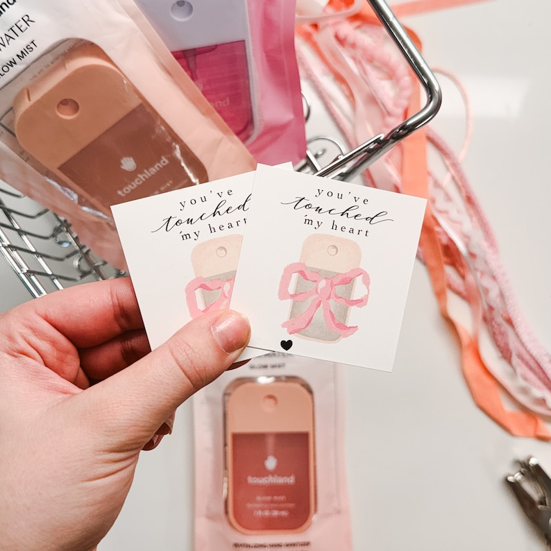 May include: Two small pink and white gift tags with the text "You've touched my heart" and a watercolor illustration of a pink bow. The tags are in a hand holding them up.
