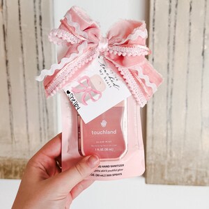 May include: A pink and white hand sanitizer bottle with a pink bow and a tag that says "You've touched my heart". The bottle is labeled "touchland Glow Mist Revitalizing hand sanitizer 1 fl oz (30 ml)" and "G Hand Sanitizer Joins skin's youthful glow 1 oz (30 ml) | 500 SPRAYS".