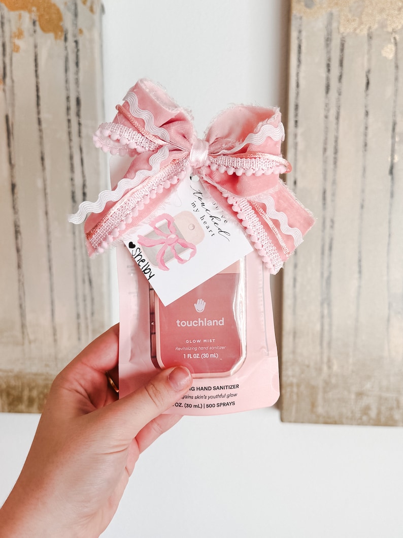 May include: A pink and white hand sanitizer bottle with a pink bow and a tag that says "You've touched my heart". The bottle is labeled "touchland Glow Mist Revitalizing hand sanitizer 1 fl oz (30 ml)" and "G Hand Sanitizer Joins skin's youthful glow 1 oz (30 ml) | 500 SPRAYS".