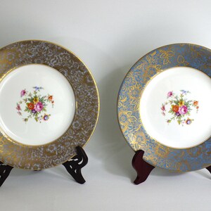 May include: Two decorative plates with floral centrepieces. One plate has a gold and grey border, the other has a blue and gold border. Both plates are displayed on dark brown stands.