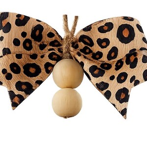 May include: A decorative bow made of tan ribbon with a black and brown leopard print pattern. The center of the bow is tied with a piece of jute twine that holds two smooth, round, light-colored wooden beads hanging vertically below the bow.