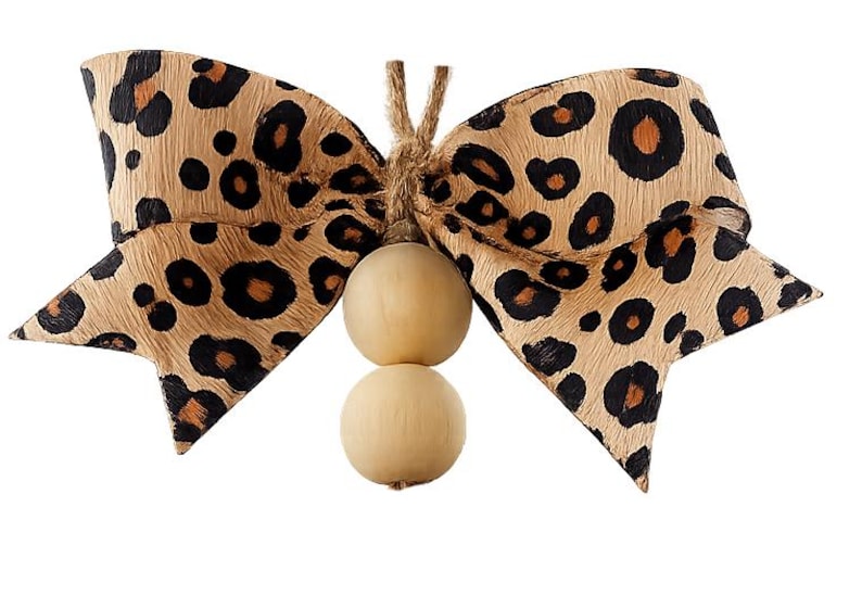 May include: A decorative bow made of tan ribbon with a black and brown leopard print pattern. The center of the bow is tied with a piece of jute twine that holds two smooth, round, light-colored wooden beads hanging vertically below the bow.