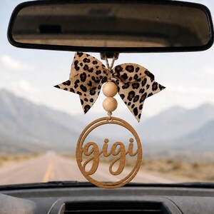 May include: A car rearview mirror hanging ornament featuring a circular wooden cutout with the name gigi in cursive script, two light-colored wooden beads, and a fabric bow with a brown and black leopard print pattern suspended by a string.
