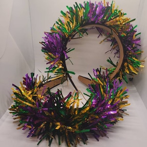 May include: Two Mardi Gras headbands with a brown base and colorful tinsel. The tinsel is in the traditional Mardi Gras colors of purple, green, and gold. The headbands are on a white surface.