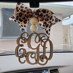 May include: A hanging car ornament featuring a brown wooden monogram with the letters EGD in a cursive script. The ornament is suspended by a jute string with two round, light-colored wooden beads and a large bow with a tan and black leopard print.