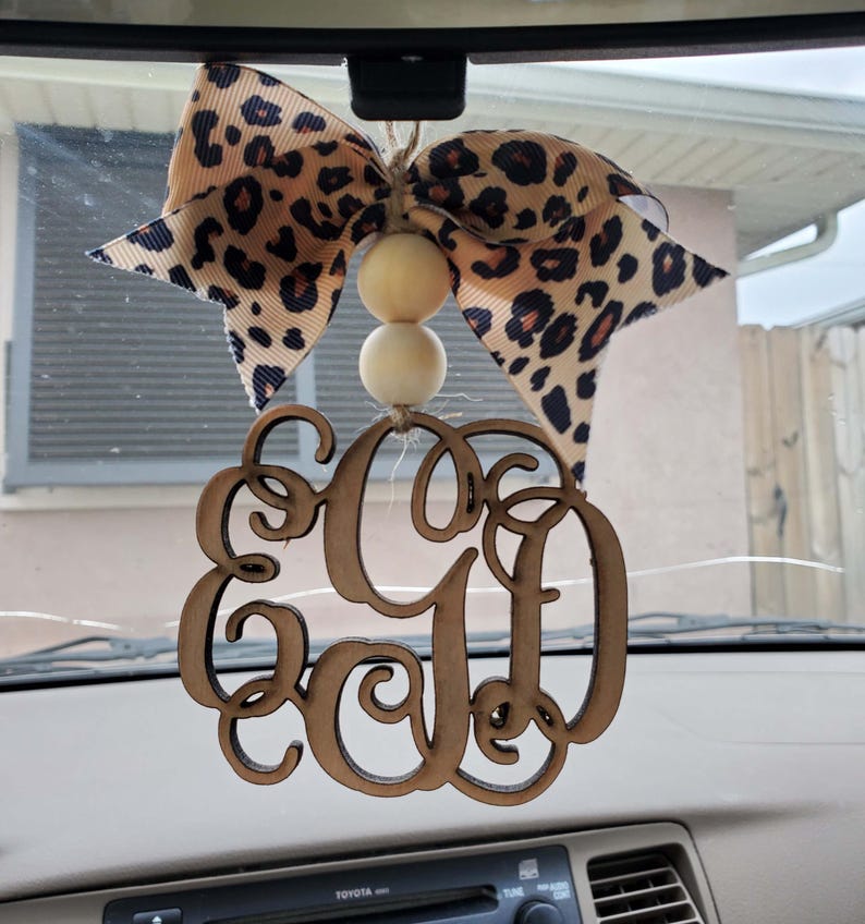 May include: A hanging car ornament featuring a brown wooden monogram with the letters EGD in a cursive script. The ornament is suspended by a jute string with two round, light-colored wooden beads and a large bow with a tan and black leopard print.