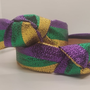 May include: A knotted headband featuring a metallic fabric with diagonal stripes in purple, green, and gold. The fabric has a shimmering, textured finish, and the headband is constructed with a central knot detail at the top, creating a wide band.