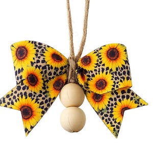 May include: A decorative bow ornament featuring a vibrant sunflower and leopard print pattern on fabric. The bow is suspended by a natural jute twine loop and accented with two smooth, round, light-colored wooden beads hanging from the center.
