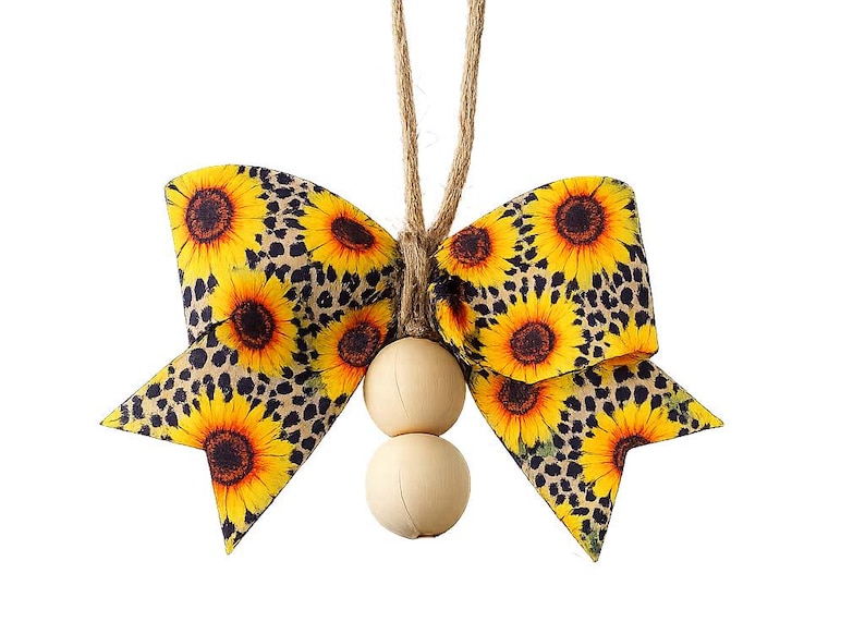 May include: A decorative bow ornament featuring a vibrant sunflower and leopard print pattern on fabric. The bow is suspended by a natural jute twine loop and accented with two smooth, round, light-colored wooden beads hanging from the center.