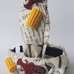 May include: Two fabric headbands with a newspaper print design. One headband has a red glitter lobster and a yellow corn cob. The other headband has a red glitter lobster and a yellow corn cob. The headbands are on a white background.