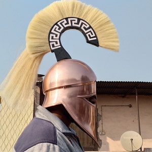 May include: A copper colored Greek warrior helmet with a white and black crest. The crest is shaped like a crescent moon with a Greek key pattern. The helmet has a face shield and a plume of white feathers.