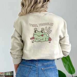 May include: A light beige sweatshirt with a green frog graphic and the text "Yeah, Toaddally" printed on the back.