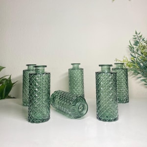 May include: Six green glass vases with a diamond pattern. The vases are cylindrical with a narrow neck and a flat base. Some vases are upright, while one is lying on its side. The vases are arranged on a white surface.