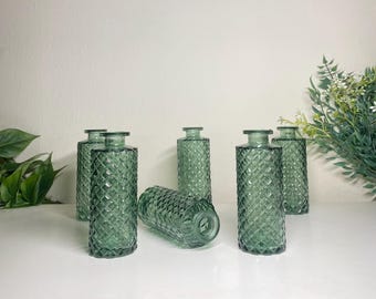 May include: Six green glass vases with a diamond pattern. The vases are cylindrical with a narrow neck and a flat base. Some vases are upright, while one is lying on its side. The vases are arranged on a white surface.