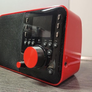 Könnte beinhalten: Ein roter und schwarzer tragbarer Lautsprecher mit einem kleinen Bildschirm und Steuertasten. Der Lautsprecher hat einen großen roten Drehregler und einen schwarzen, strukturierten Lautsprechergrill. Das Gerät ist für die Audiowiedergabe konzipiert.