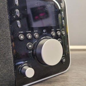 Pode incluir: Sistema de música digital Logitech preto e prata com tela e botões de controle. O dispositivo possui um mostrador central grande e um mostrador de volume menor. O alto-falante é coberto com um tecido preto texturizado.