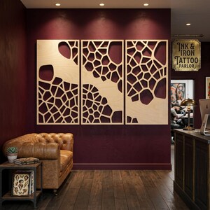 May include: Three wooden wall panels with a geometric, openwork design are mounted on a deep red wall. The panels are made of light-coloured wood and feature a pattern of irregular shapes. The scene includes a leather armchair and a small side table.