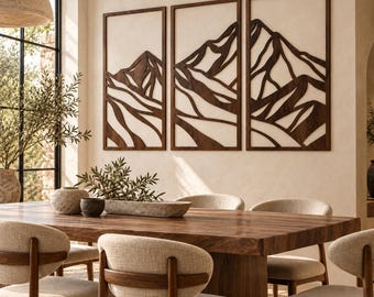 May include: A set of three rectangular dark brown wooden wall art panels featuring a cutout mountain range design. Each panel is framed in a thin wood border, displaying a layered, abstract landscape of peaks and valleys mounted on a wall.