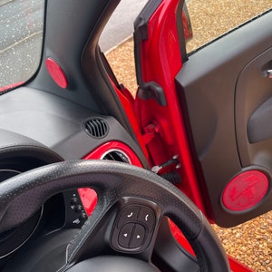 Può includere: Primo piano dell'interno di un'auto rossa. Il volante è nero con una finitura argentata e ha pulsanti per controllare il telefono e il sistema audio. La portiera dell'auto è aperta e l'interno è nero.
