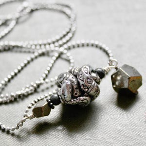 May include: A silver chain necklace with a silver pendant featuring a detailed snake design. The pendant is accented with a small, dark grey stone and a larger, grey, faceted stone.