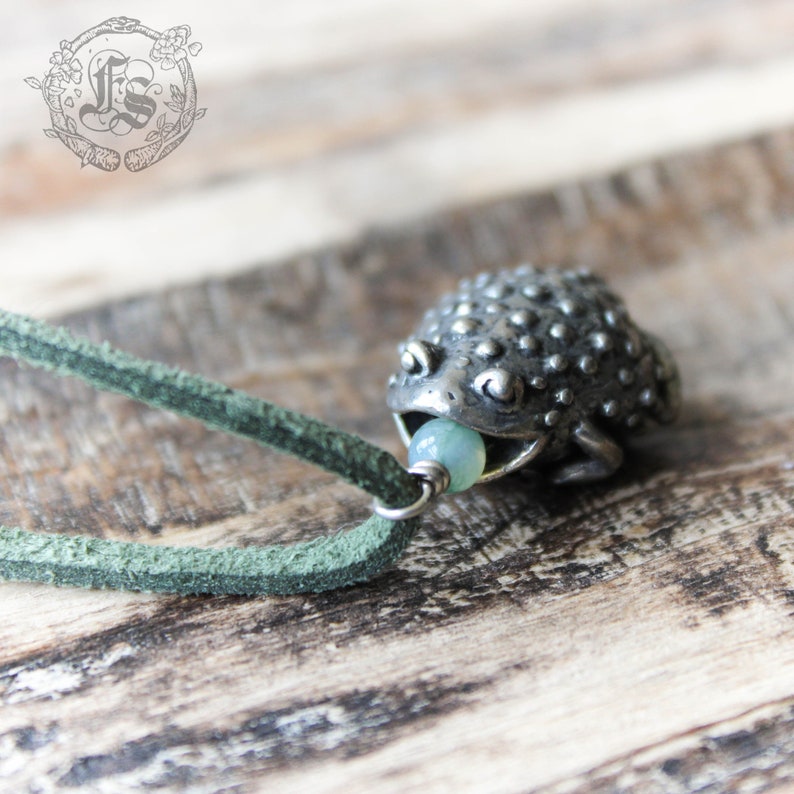 May include: A silver frog pendant with a light blue bead in its mouth, hanging from a green leather cord.