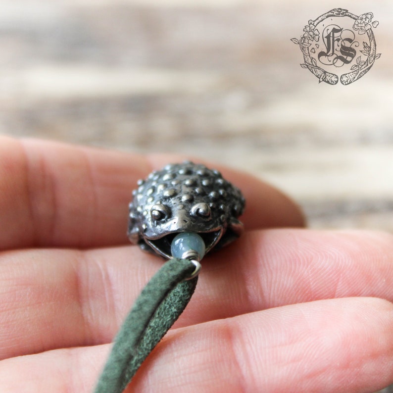 May include: A silver toad charm with a light blue bead in its mouth, hanging from a green suede cord.