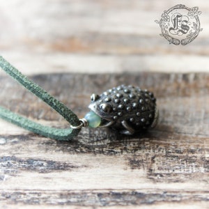 May include: A silver frog pendant with a green stone in its mouth, hanging from a green leather cord.