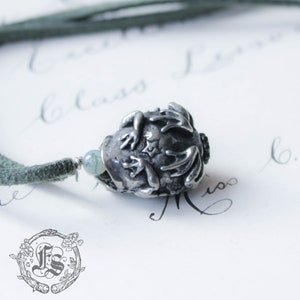 May include: A silver charm necklace with a frog pendant. The frog is curled up with its legs tucked in. The necklace is on a green cord.