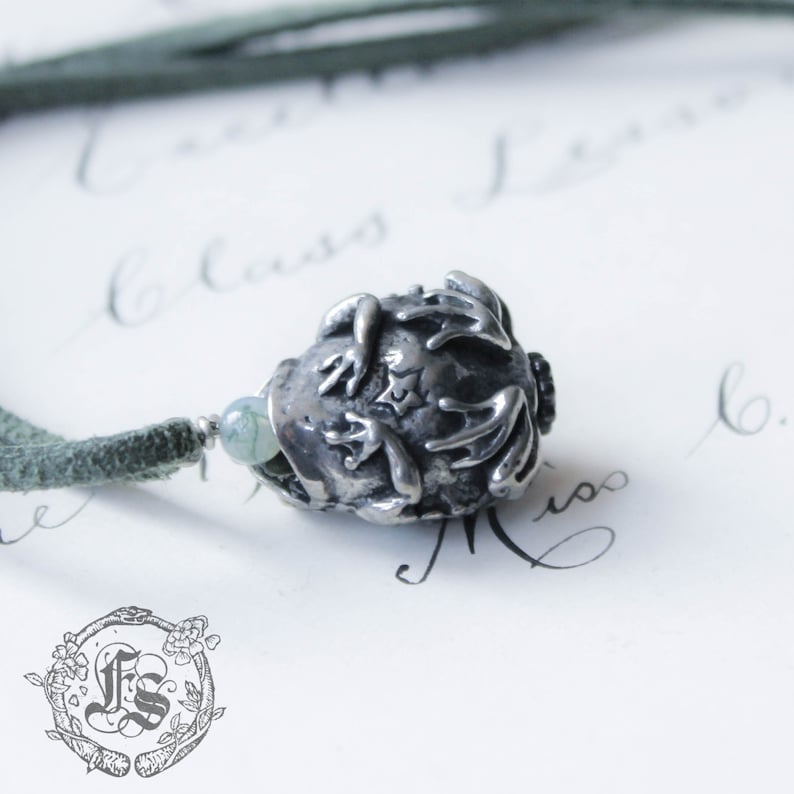 May include: A silver charm necklace with a frog pendant. The frog is curled up with its legs tucked in. The necklace is on a green cord.