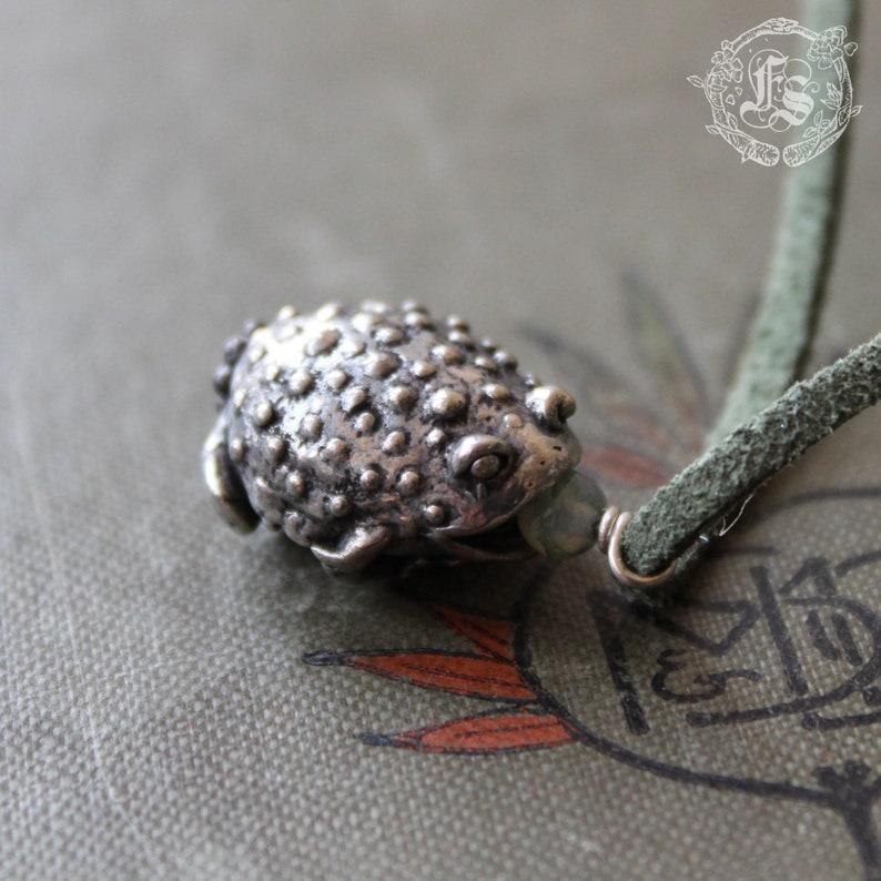 May include: A silver toad charm necklace with a green stone bead. The toad is detailed with textured bumps and has a realistic appearance.