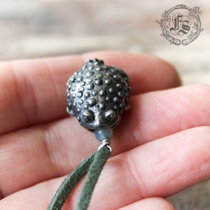 May include: A silver toad charm with a green suede cord. The toad is detailed with a bumpy texture and has a bead below it.