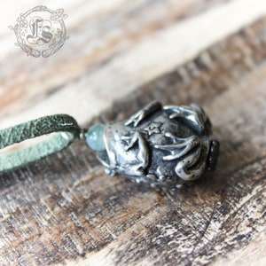 May include: A silver pendant featuring a detailed frog design with a green stone in its mouth. The pendant is attached to a green suede cord.