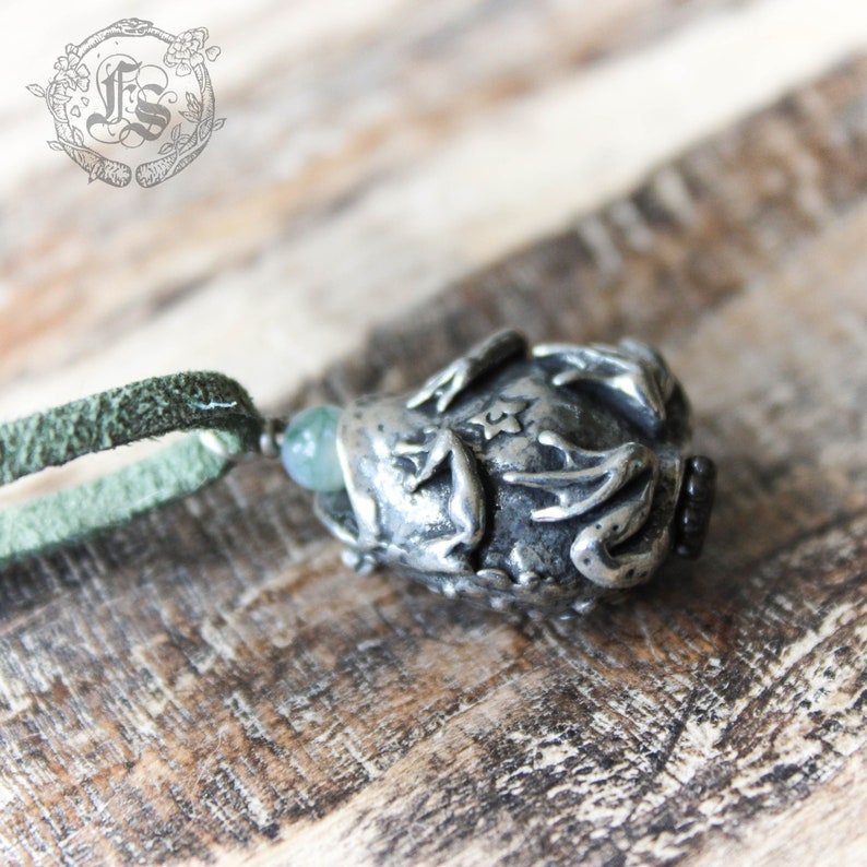 May include: A silver pendant featuring a detailed frog design with a green stone in its mouth. The pendant is attached to a green suede cord.