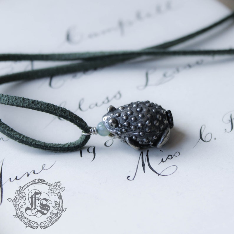 May include: A silver toad pendant with a green gemstone bead on a dark green suede cord.