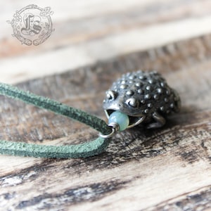 May include: A silver frog pendant with a light blue bead in its mouth, hanging from a green leather cord.