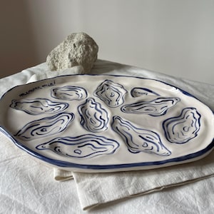 Op de afbeelding: Ovale keramische oesterserveerschaal met een witte basis en een donkerblauwe rand. De schaal heeft handgeschilderde oesterschelpontwerpen in blauw en de Italiaanse uitdrukking "mamma mia!" staat op de rand.