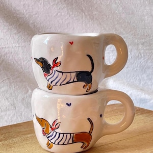 Può includere: Due tazze in ceramica bianca impilate. Ogni tazza presenta un disegno di bassotto dipinto a mano, con una sciarpa rossa e una camicia a righe blu e bianche. Piccoli cuori rossi e blu sono sparsi intorno alle tazze.