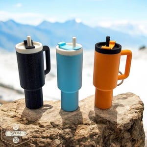 May include: Three insulated travel mugs in black, blue, and orange, each with a handle and a lid with a straw. The mugs are displayed on a rock with a blurred mountain backdrop. The mugs are designed to keep drinks cold or hot.
