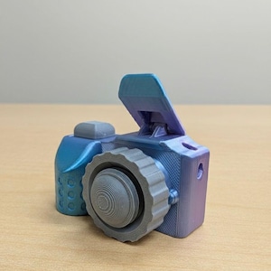 以下が含まれることがあります： 青と紫の色合いの3Dプリントされたおもちゃのカメラ。グレーのレンズと小さな青いアクセサリーが付いています。カメラは、ビューファインダーやテクスチャードグリップなど、詳細なデザインになっています。
