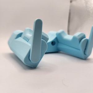 Puede incluir: Un soporte para teléfono plegable de plástico azul claro. El soporte tiene una superficie texturizada y un borde elevado para sujetar un teléfono en su lugar.