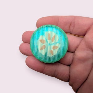 May include: A round, wooden teal-colored button with a paw print design. The paw print is composed of four beige-colored pads. The button has a textured, ridged edge.