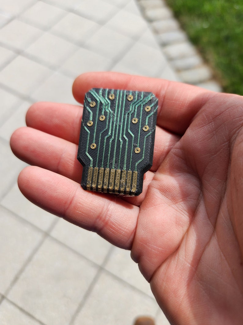 以下が含まれることがあります： 緑色のトレースと金色のピンが付いた黒い回路基板のクローズアップ。基板は小さく長方形で、多数の小さな穴があります。