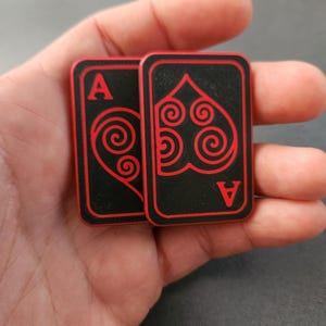Può includere: Due carte da gioco nere con bordi rossi e disegni di cuori. Le carte presentano la lettera "A" in rosso in alto e in basso. Il disegno del cuore è un motivo a spirale stilizzato.