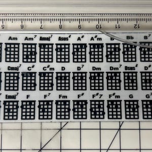 May include: A white plastic ukulele chord chart with black text and diagrams. The chart includes chords for A, Am, Asus, Bb, B, Bm, C, Cm, D, Dm, E, Eb, F, Fm, G, Gm, and more.