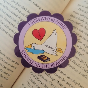 Può includere: Un adesivo viola e giallo con un'oca bianca, un cuore spezzato e un libro. L'adesivo dice "I Survived Reading Sunrise on the Reaping".